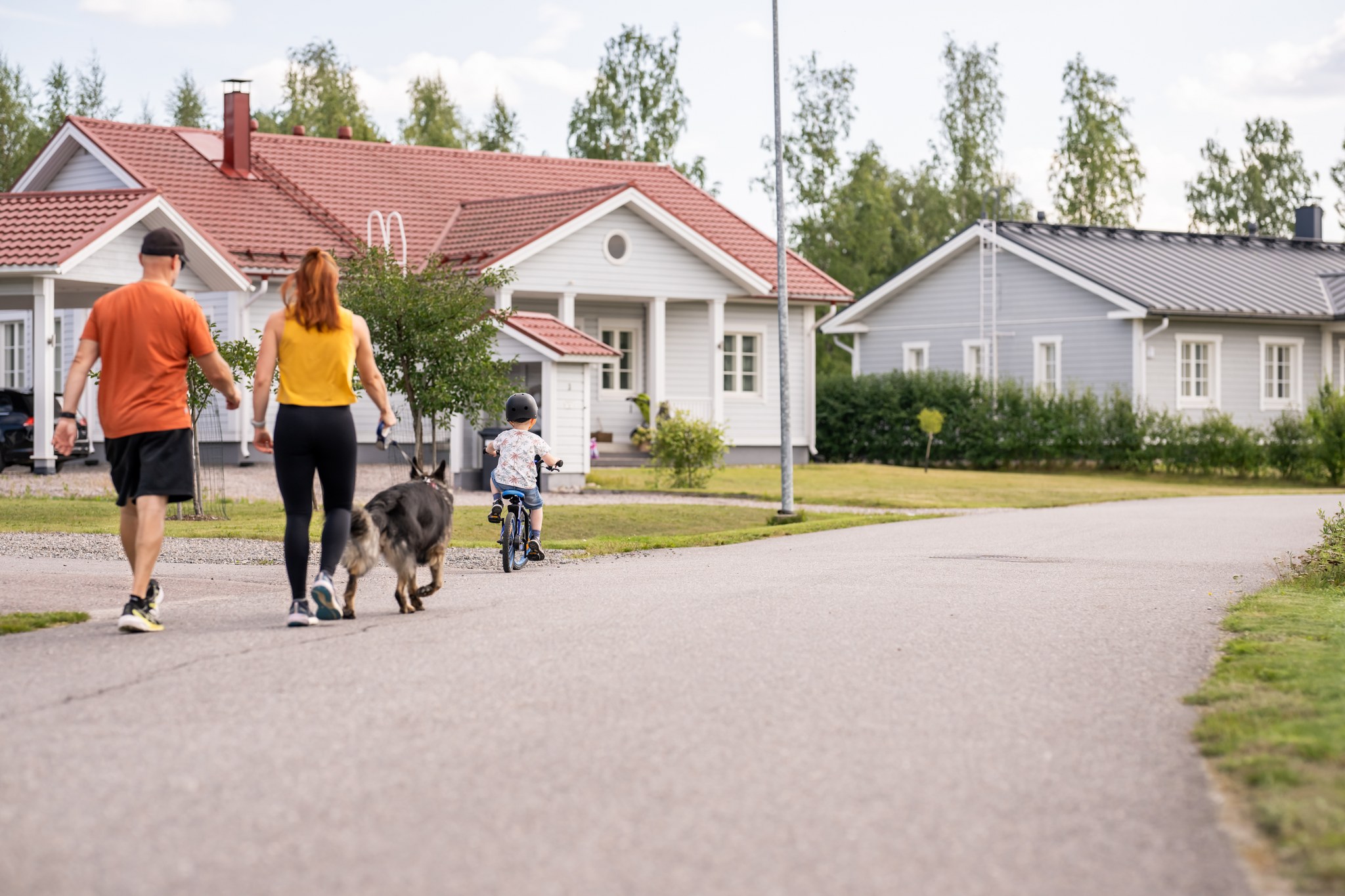 Perhe, äiti, isä ja lapsi sekä koira kulkevat tietä pitkin asuntoalueella.